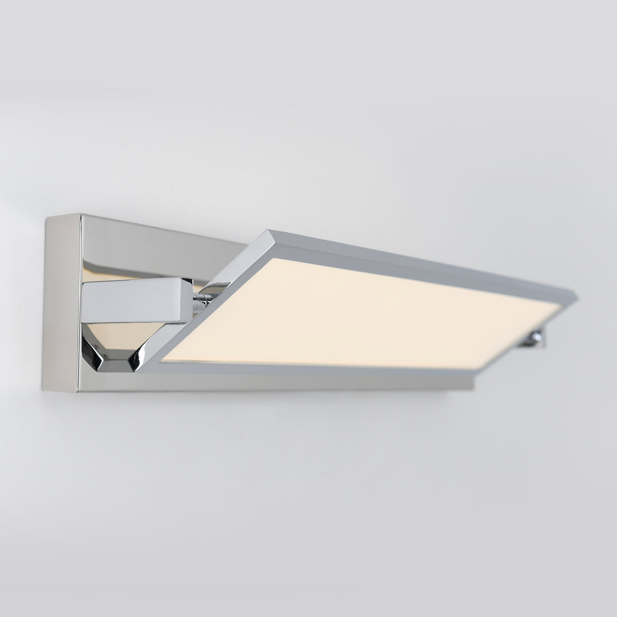 A modern, rectangular wall-mounted LED light fixture with a chrome finish and a flat, angled panel emitting soft white light, set against a plain light gray background.