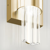 A modern wall sconce with a gold rectangular base and cylindrical clear glass rods, emitting soft light against a white wall. The design features a blend of metallic and transparent elements.