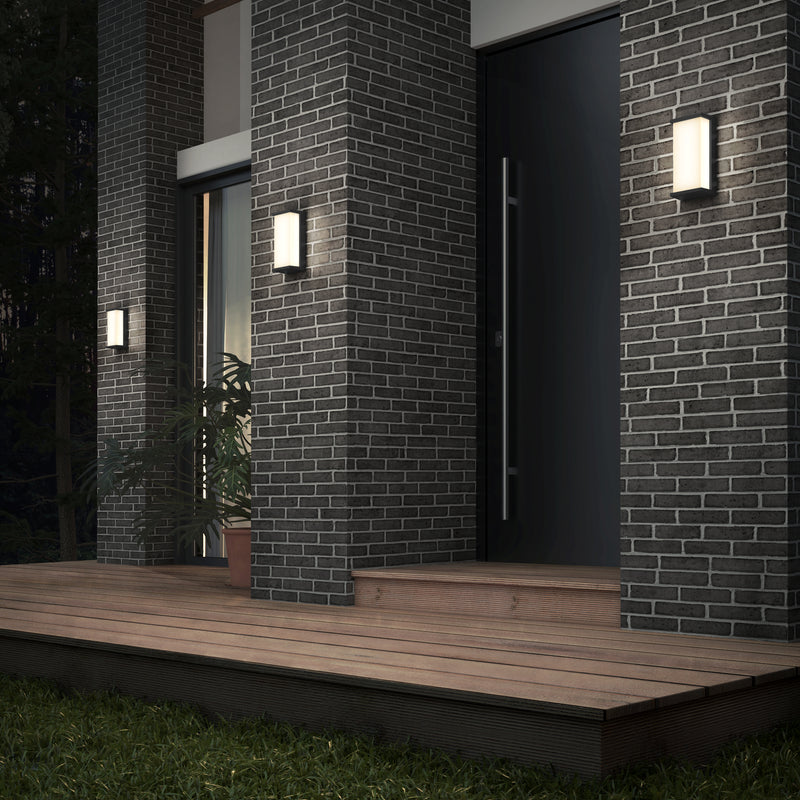 A modern entrance features dark brick walls lit by three Artika Faro modern LED indoor-outdoor wall lights (3 CCT). A wooden deck leads to a black door, with a potted plant near the left window and grass bordering the decking.