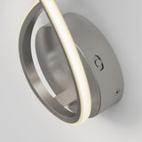 A modern, circular LED wall sconce with a metallic finish is mounted on a light-colored wall. The fixture features a sleek, curved design with a glowing light strip along its edge.