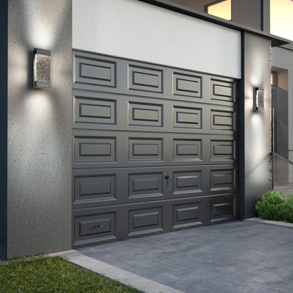 A modern house with a closed dark gray garage door featuring rectangular panels. On either side are Artika Essence Glow Box indoor-outdoor LED wall lights, with steps and green bushes to the right.