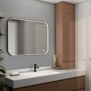 A modern bathroom features the Artika Royce Dimmable LED Mirror (3 CCT, mounts horizontally or vertically) above a white sink with a black faucet, wooden cabinets, a reed diffuser, and a small plant. Light enters through window blinds.