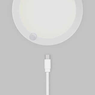 A white USB-C charging cable lies below the Artika Accent under cabinet LED light 3CCT, a round white device with a small circular sensor on its edge, all placed on a light gray surface.