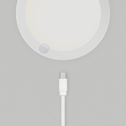 A white USB-C charging cable lies below the Artika Accent under cabinet LED light 3CCT, a round white device with a small circular sensor on its edge, all placed on a light gray surface.
