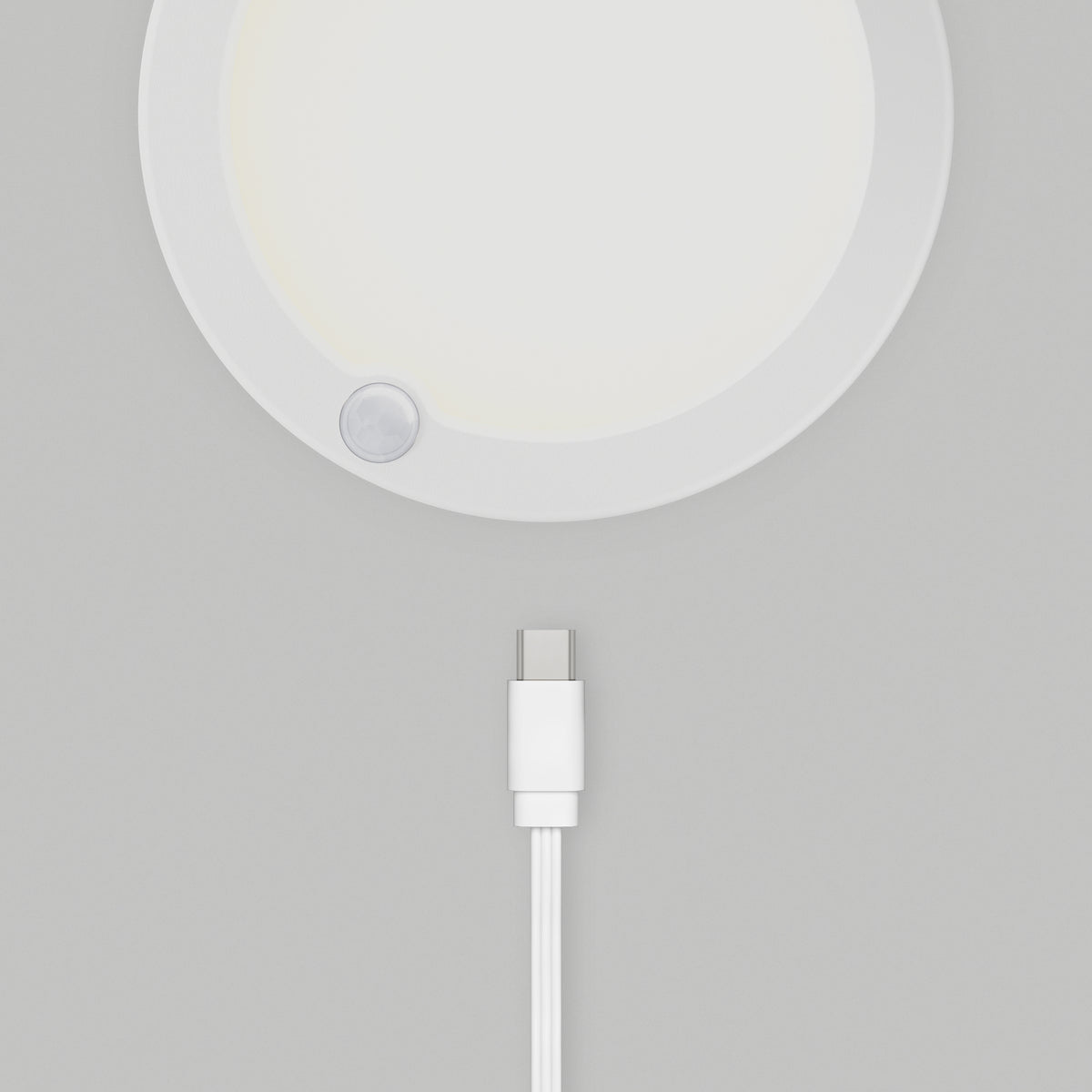 A white USB-C charging cable positioned below a round, white electronic device mounted on a light gray wall. The device has a small circular sensor or button near its edge.