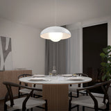 A modern dining room with a round table set for four, black chairs, a large white pendant lamp overhead, and a window showing a night view. A plant and wooden sideboard are in the background.