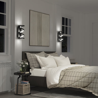 A modern bedroom features a neatly made bed with plaid bedding, two large pillows, and a throw blanket. Swirl indoor-outdoor LED wall lights by Artika illuminate the space. A black side table with books and a plant sits beside the bed.