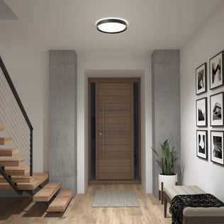 A modern entryway with a wooden door, floating stairs with metal railings, a potted plant, bench, black-and-white art, wooden floors, and an Artika Callisto 15 in. LED flush mount light (5 CCT) on the ceiling.