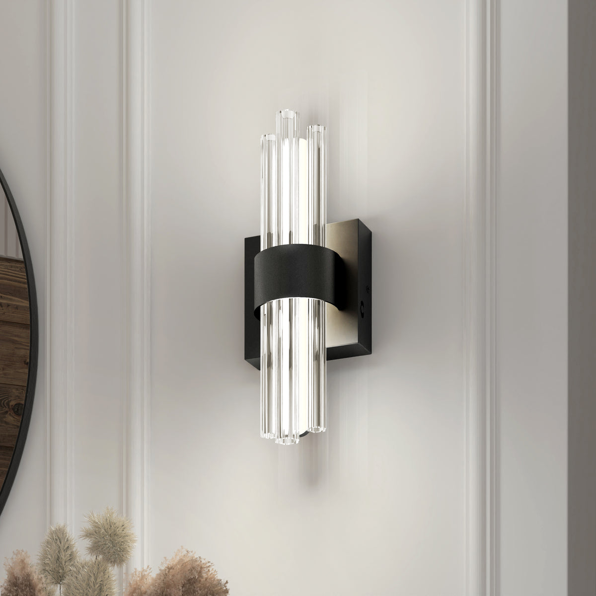 A modern wall sconce with vertical clear glass rods and a black metal band is mounted on a light-colored paneled wall. Dried flowers are visible in the bottom left corner of the image.
