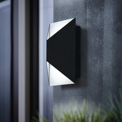 The Artika Tria LED indoor-outdoor wall light, featuring a modern matte black geometric design, is mounted on a textured gray wall and emits soft LED lighting. Blurred windows and plants appear in the background.