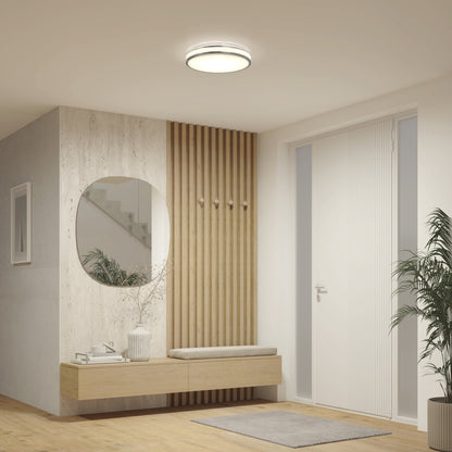 Modern entryway with light wood bench, textured wall paneling, round mirror, hooks, potted plant, and minimalist decor. Features Artika's Hemisphere 15 in. LED flush mount light 5 CCT for adjustable brightness, plus white walls and a door with vertical blinds.
