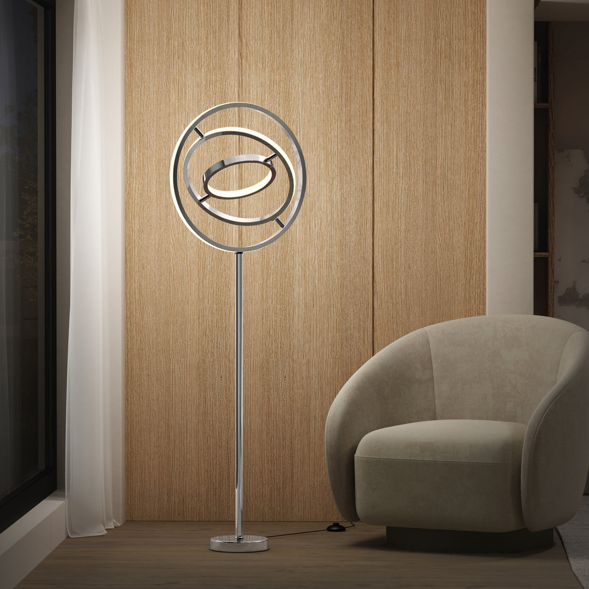 The Artika Pivot modern LED floor lamp 3 CCT, featuring circular rings, stands beside a curved beige armchair in a minimalist room with wooden paneling and a sheer white curtain by the window.