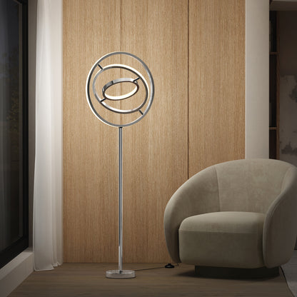 The Artika Pivot modern LED floor lamp 3 CCT, featuring circular rings, stands beside a curved beige armchair in a minimalist room with wooden paneling and a sheer white curtain by the window.