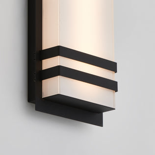 A close-up of the Artika Glowbox 3 indoor-outdoor LED wall light, featuring a frosted glass shade with black metal horizontal bands and a black rectangular backplate, mounted on a light-colored wall.