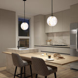 A modern kitchen with beige cabinets, marble countertops, and two pendant lights above a wooden island. Two cushioned chairs face the island, which has bread, cheese, and a glass of coffee set on it.