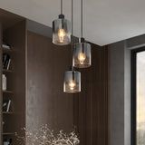 Three modern pendant lights with transparent cylindrical glass shades hang at different heights in a stylish, contemporary room featuring wooden paneling, a bookshelf, and a large window.