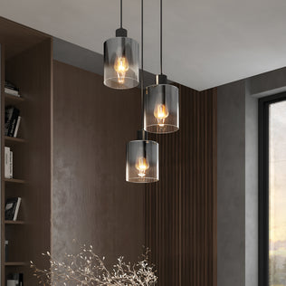 The Artika Haze modern 3-light pendant light with cylindrical glass shades hangs in a contemporary room featuring wood paneling, a built-in bookshelf, and a large window allowing abundant natural light.