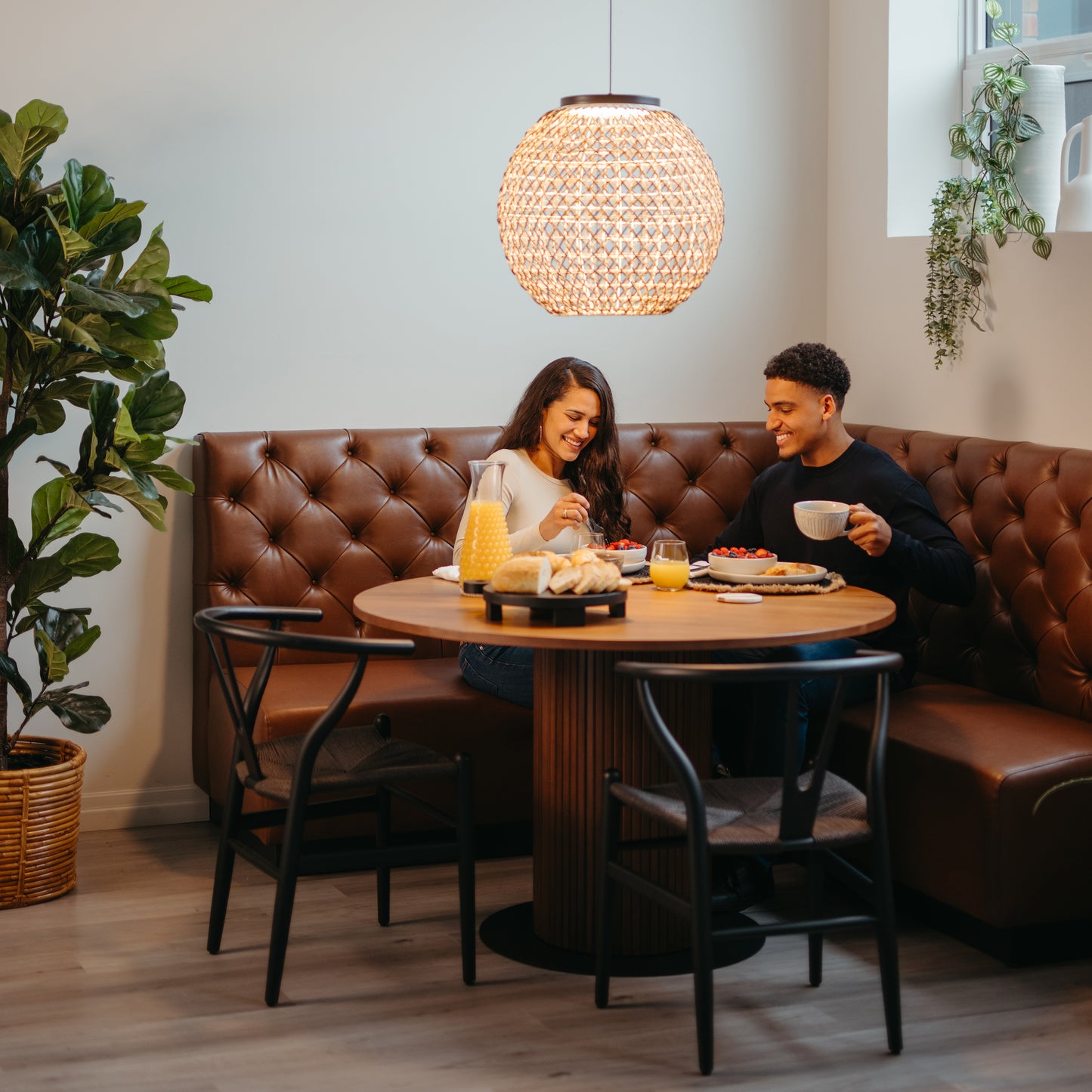 A woman and man sit at a round table in a cozy cafe booth, enjoying breakfast with pastries, orange juice, and coffee. The warm glow from the Artika Benson LED pendant light 5 CCT adds to the inviting atmosphere filled with plants.
