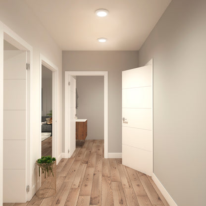 A modern hallway features light gray walls, wood flooring, white doors, and an Artika Europa Disk 7 in. LED modern flush mount light overhead. A small green plant in a basket sits on the floor near an open door to a bathroom with a wooden vanity.