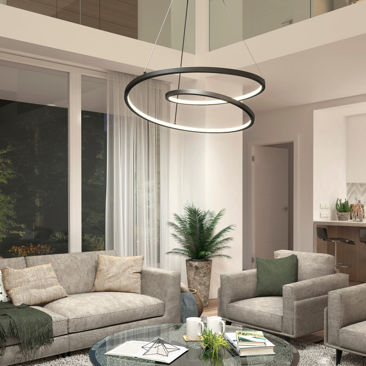 Modern living room with two gray sofas, an armchair, and a glass coffee table. Large windows, sheer white curtains, a potted plant, and a circular, modern chandelier hanging from the ceiling. Nighttime view outside.