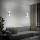 A modern living room with a gray sectional sofa, brown and gray cushions, wall-mounted sconces, white paneled walls, a marble coffee table, a plant, and large windows with beige curtains showing a city view at night.