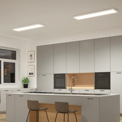 Modern kitchen with light gray cabinets, a central island with two brown chairs, built-in ovens, under-cabinet lighting, a potted plant by the window, and two Artika Skyshade smart RGB LED light panels on the ceiling.