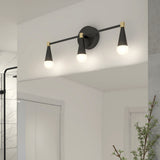 A modern bathroom light fixture with three black and gold cone-shaped lights is mounted above a mirror, illuminating the white wall and reflecting in the glass. A shower and a plant are partially visible nearby.