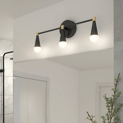 The Artika Wayden LED mid-century bathroom vanity light (3 CCT) features black and gold finishes with three cone-shaped shades above a mirror, casting warm light. Part of a glass shower and a leafy plant are also in view.