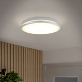 A modern round ceiling light fixture emits a soft, even glow; part of a wooden slat wall, a green plant, and a gray door are visible beneath the white ceiling.