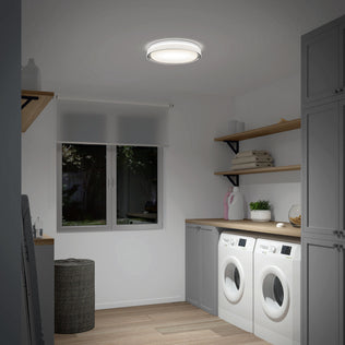 Modern laundry room with front-loading washer and dryer, gray cabinets, wooden shelves holding towels, a laundry basket, a window with a roller shade, and the Artika Niko 15.5 in. modern LED flush mount light 5 CCT on the ceiling.