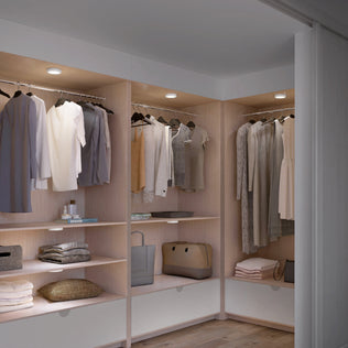 A modern walk-in closet with neatly hung clothes, folded garments, bags, and accessories. The space is well-lit by Artikas Accent under cabinet LED light 3CCT and features wooden shelves and white drawers.