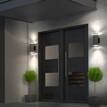 Modern double front doors with frosted glass panels, framed by round potted shrubs. Two Artika Trygon indoor-outdoor wall lights illuminate the doors and gray concrete exterior, highlighting the sleek, contemporary entrance.