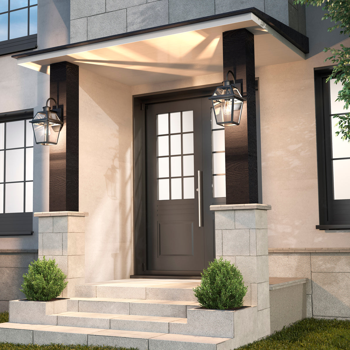 A modern house entrance with a dark front door, glass panels, two large lantern-style wall lights, stone steps, and small green shrubs on either side of the doorway.