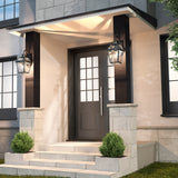 A modern house entrance with a dark front door, glass panels, two large lantern-style wall lights, stone steps, and small green shrubs on either side of the doorway.