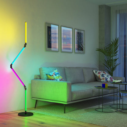 A modern living room with a gray sofa, colorful pillows, an Artika Fitch smart foldable RGB LED gaming floor lamp, framed art, a potted plant, and a coffee table with a Rubik’s cube on a rug.