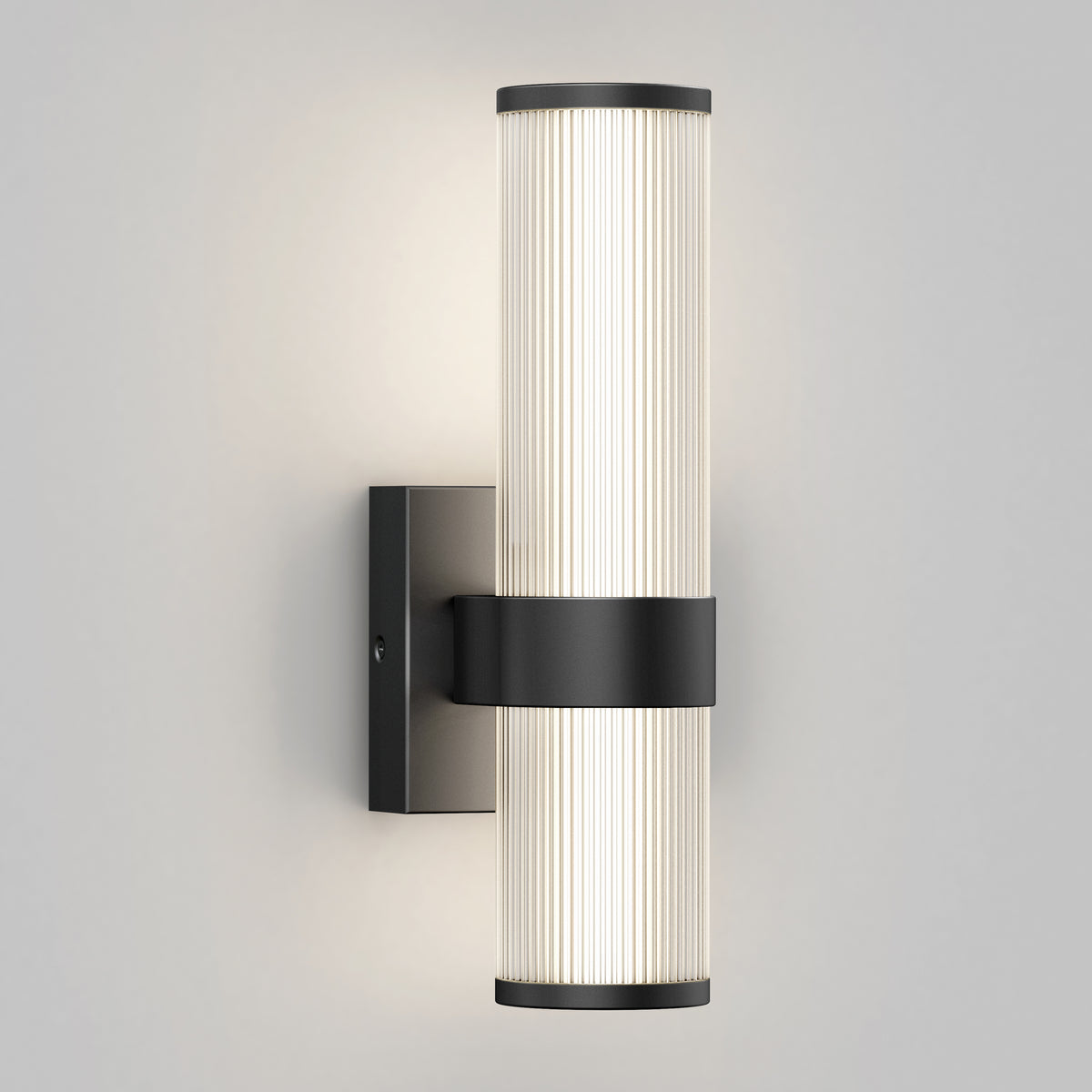 A modern wall sconce with two vertically aligned cylindrical frosted glass shades, mounted on a black rectangular base, emitting a soft, even light against a plain light gray wall.