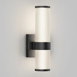 A modern wall sconce with two vertically aligned cylindrical frosted glass shades, mounted on a black rectangular base, emitting a soft, even light against a plain light gray wall.