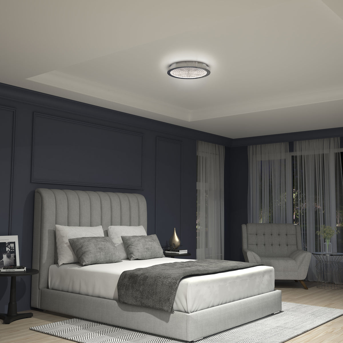 A modern bedroom with dark blue walls, a large upholstered bed with gray and white bedding, a round ceiling light, a side table with decor, a gray armchair by the window, and natural light coming through sheer curtains.