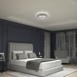 A modern bedroom with dark blue walls, a large upholstered bed with gray and white bedding, a round ceiling light, a side table with decor, a gray armchair by the window, and natural light coming through sheer curtains.