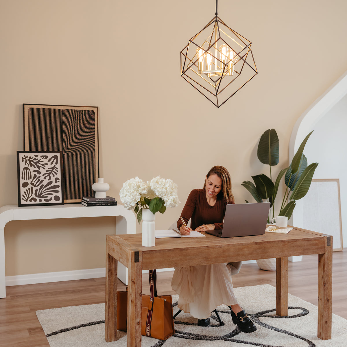 A woman sits at a wooden desk in a stylish, modern home office, writing in a notebook beside a laptop, a vase of white flowers, and décor, with plants and artwork in the background.