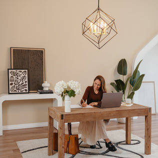 A woman sits at a wooden desk with a laptop, writing in a notebook. The workspace features plants, artwork, and an Artika Harmonium modern mid-century pendant light for a cozy, stylish atmosphere.