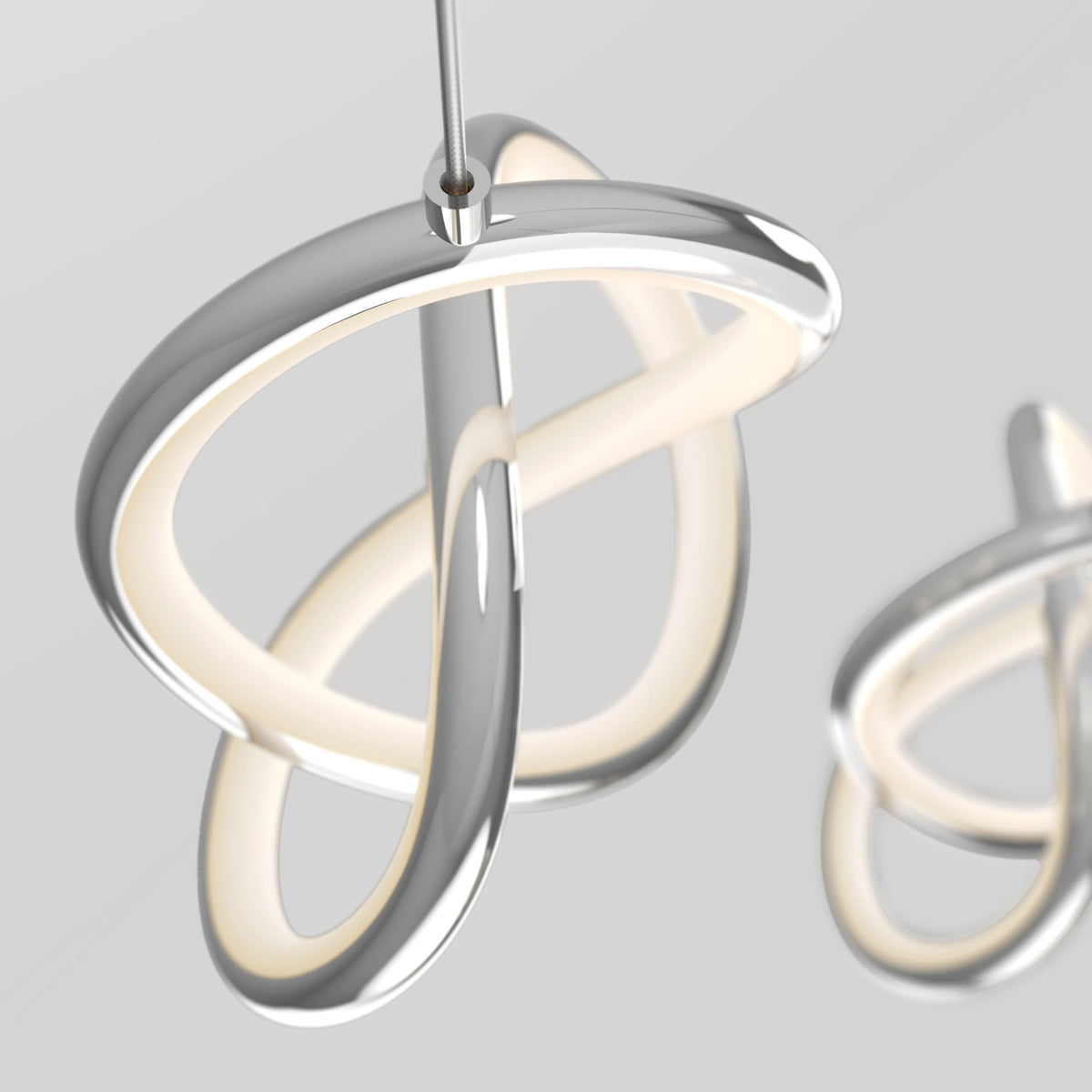 A modern, sculptural LED pendant light with smooth, intertwined silver loops and a soft inner glow, hanging from a thin cable against a neutral gray background. Another similar light is blurred in the background.