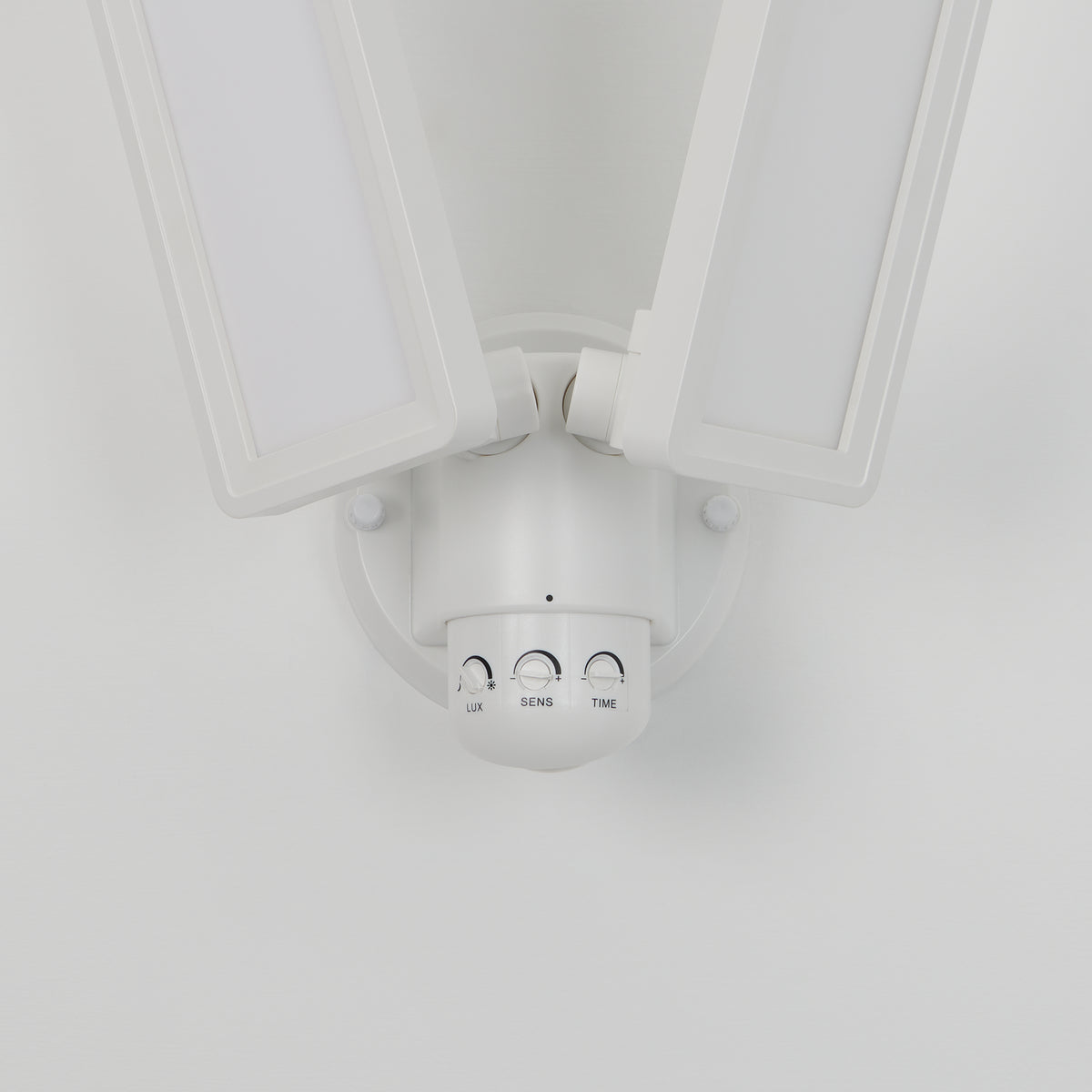 A close-up view of a modern white double LED security light fixture, mounted on a wall, with adjustable panels and three control dials labeled LUX, SENS, and TIME.