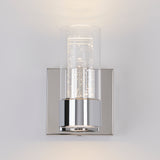 A modern wall sconce with a chrome base and a cylindrical glass shade, featuring small LED lights inside that create a sparkling effect against a light gray wall.