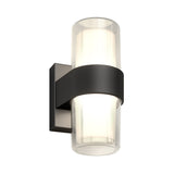 A modern wall sconce with a cylindrical frosted glass shade, partially encased by a horizontal black metal band, mounted on a square black backplate, emitting a soft white light.