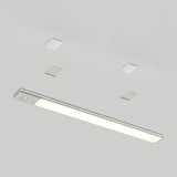 A slim, rectangular LED ceiling light fixture with a metallic finish is installed on a white ceiling, accompanied by four small square-shaped ceiling elements in two rows above it.