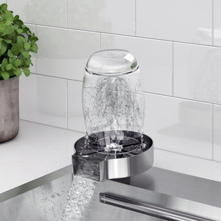 A glass cup is placed upside down on a glass rinser beside the Artika Neoflow 31 in. stainless steel workstation sink, with water spraying upwards to clean the inside, next to a potted plant and white tiled backsplash.