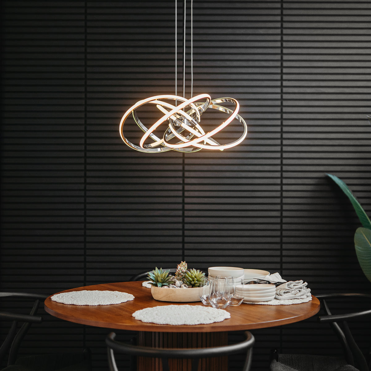 A modern dining area with a round wooden table set for four, featuring placemats, plates, and glasses. Above the table hangs a contemporary LED pendant light with interlocking rings, against a dark, textured wall.