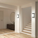 Minimalist hallway with light wooden floor, white walls, a staircase on the right, a dark wood console table with flowers and books on the left, and a large round mirror above the table. Modern wall sconces provide lighting.