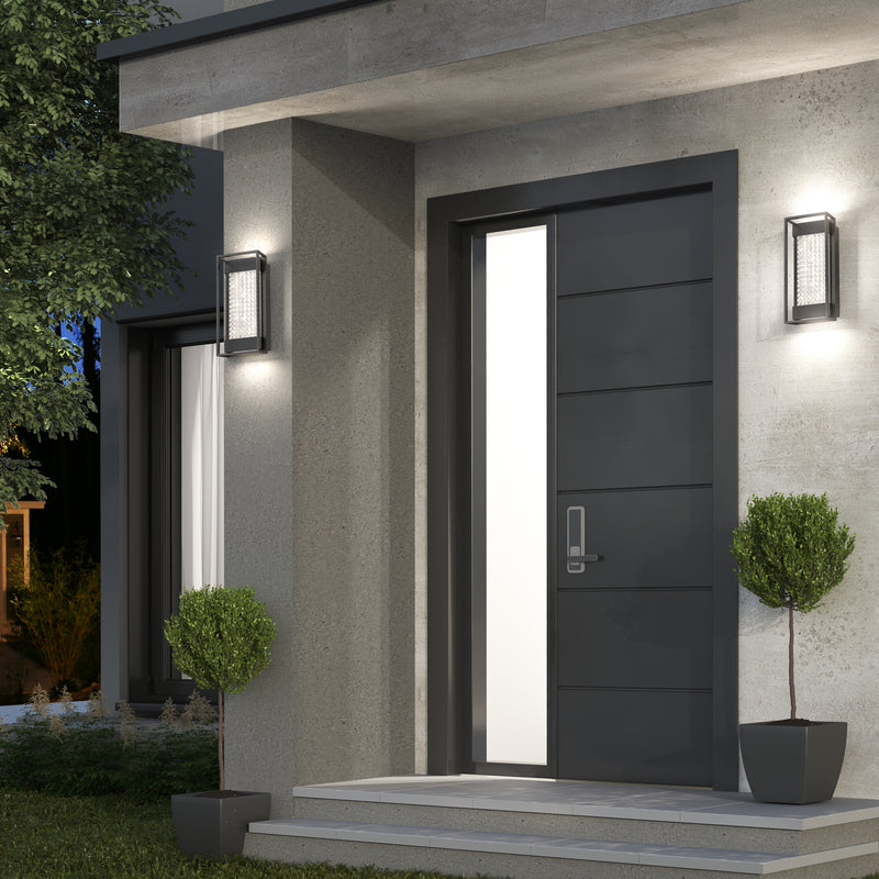 Modern front entrance with a sleek black door, vertical frosted glass panel, silver handle, and two potted topiary plants. Artika Glam indoor-outdoor LED wall lights are mounted on each side against concrete walls by the small porch.